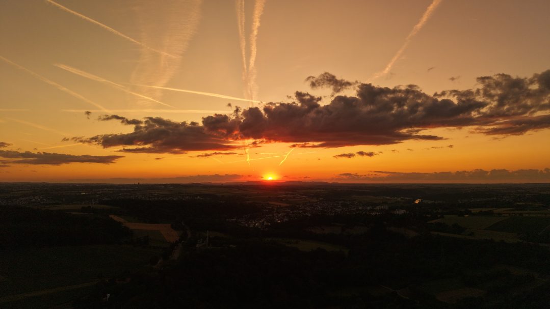 Sonnenuntergang über Hegnach – die Sonne sinkt als verglühender Punkt am Horizont, während zahlreiche Kondensstreifen kreuz und quer den Abe